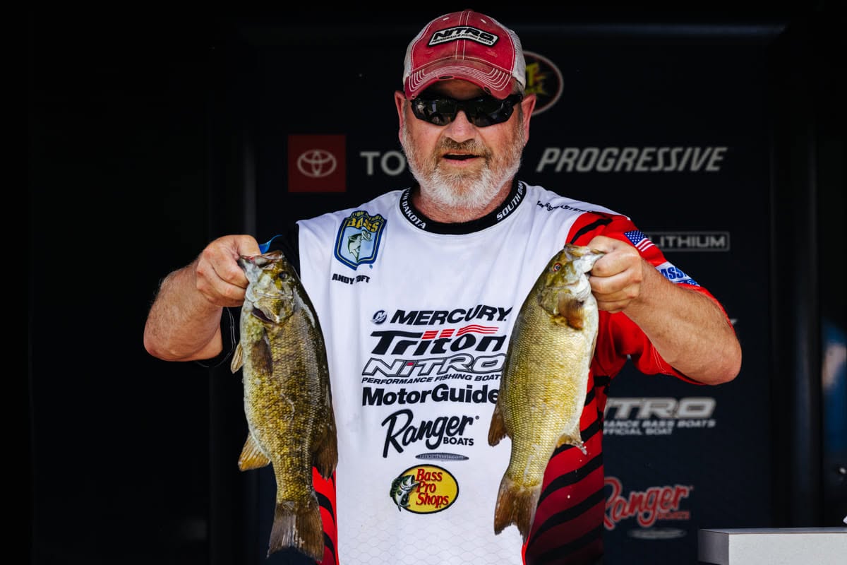 Angler in a red cap holding up two smallmouth bass at a tournament weigh-in