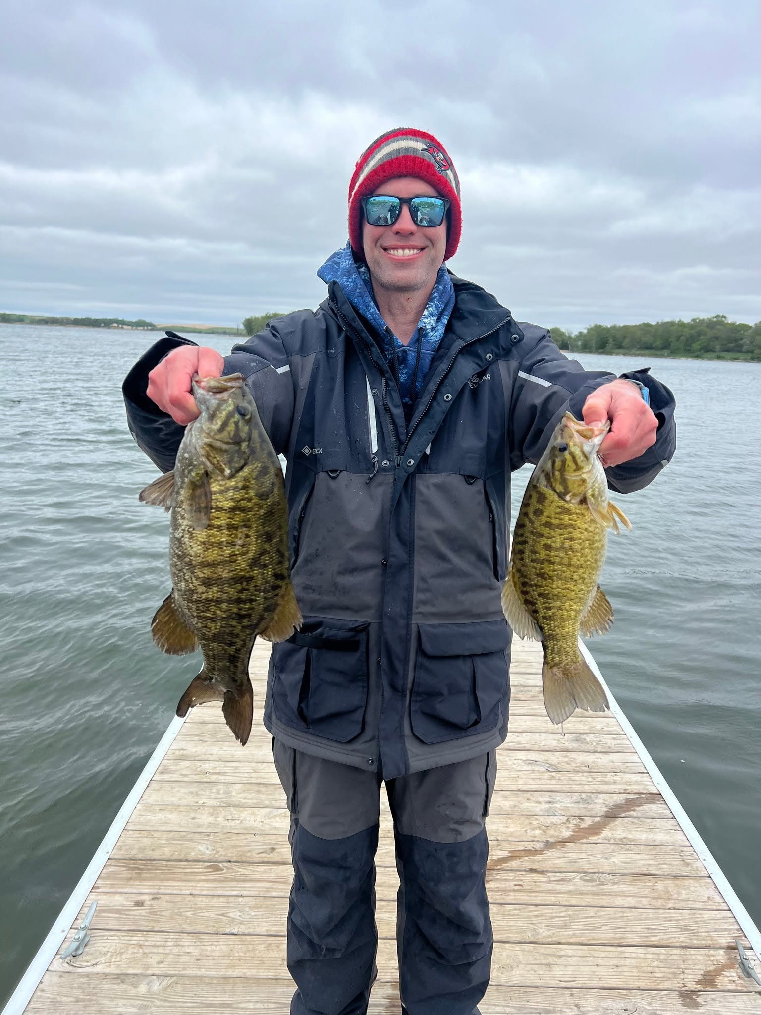 Angler in cold-weather gear holding two smallmouth bass on a dock