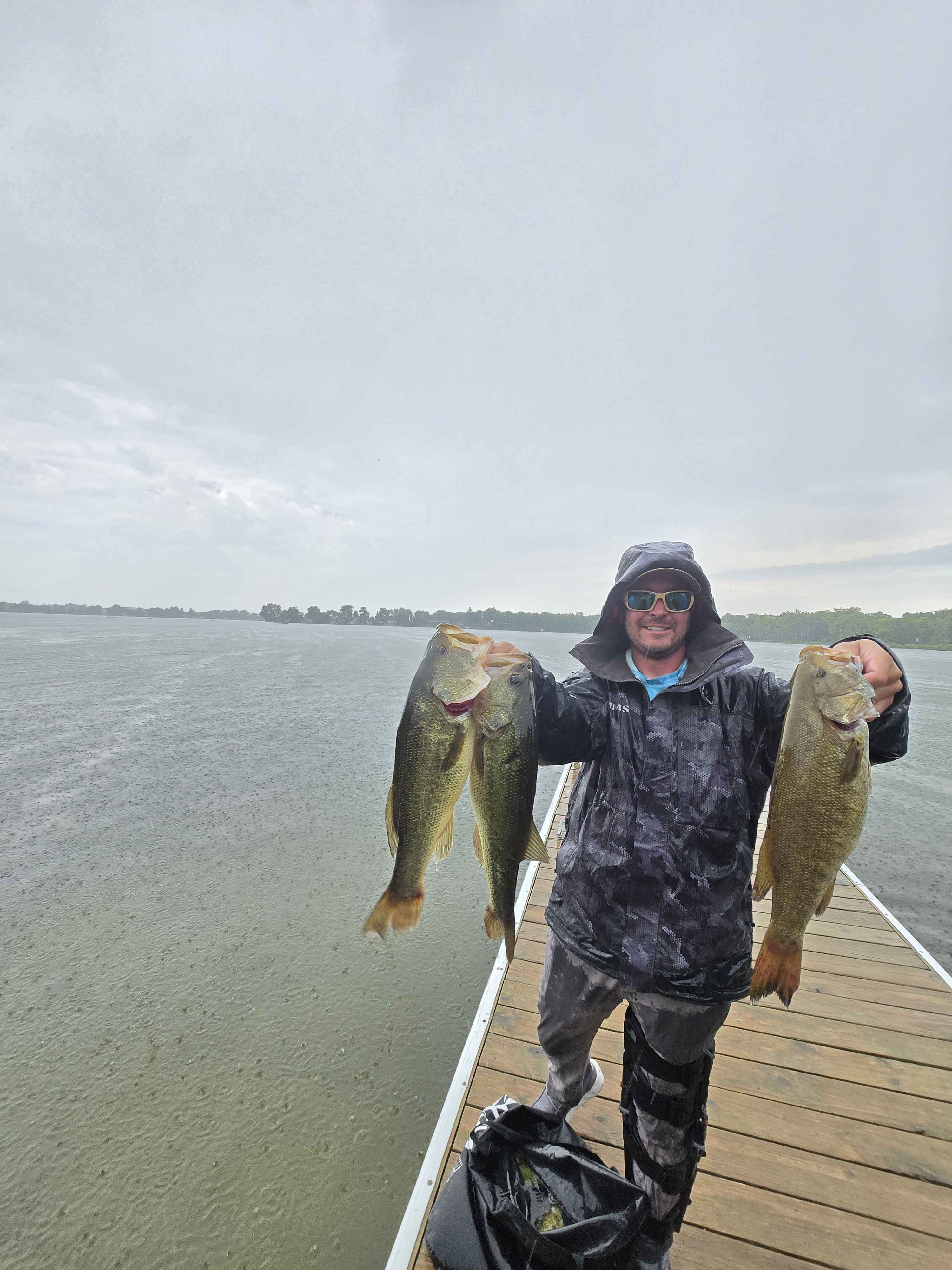 Angler in rain gear holding two largemouth and a smallmouth bass on the Enemy Swim dock